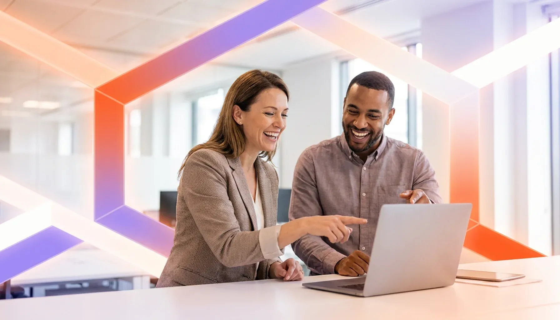 Two smiling colleagues pointing at a laptop screen in a modern office.