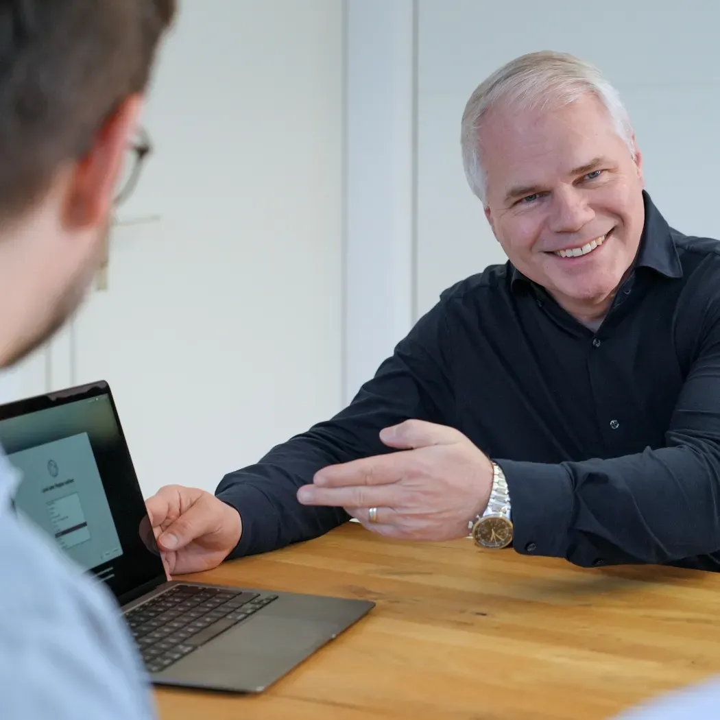 A smiling man points to a colleague's laptop during a discussion at a wooden table.
