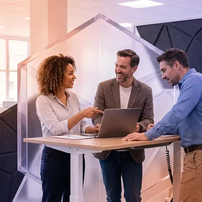 Three colleagues smiling and collaborating around a laptop at a standing desk.