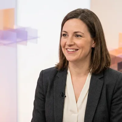 Smiling woman wearing a dark blazer and lapel microphone during a studio interview.