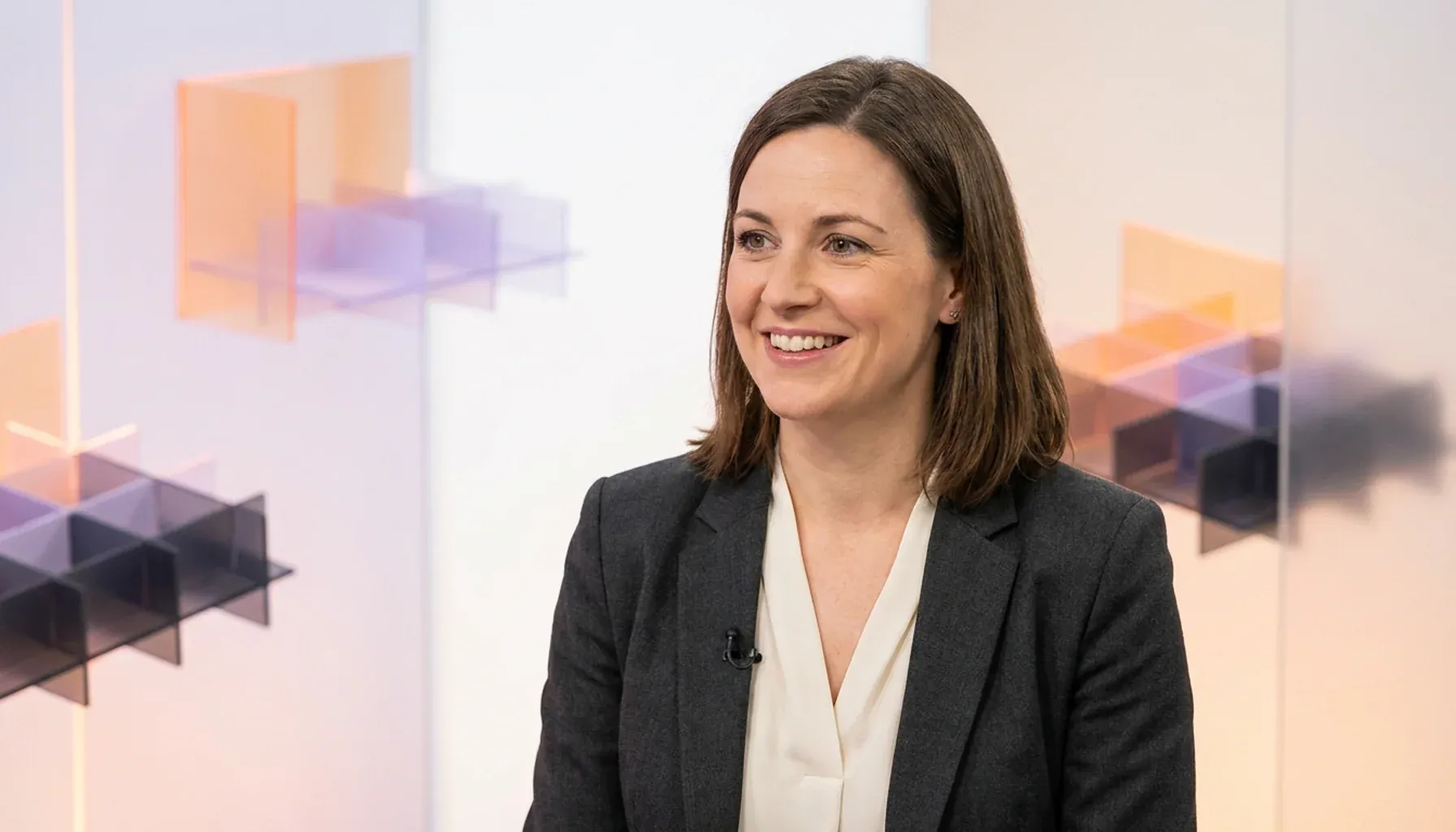 Smiling woman wearing a dark blazer and lapel microphone during a studio interview.
