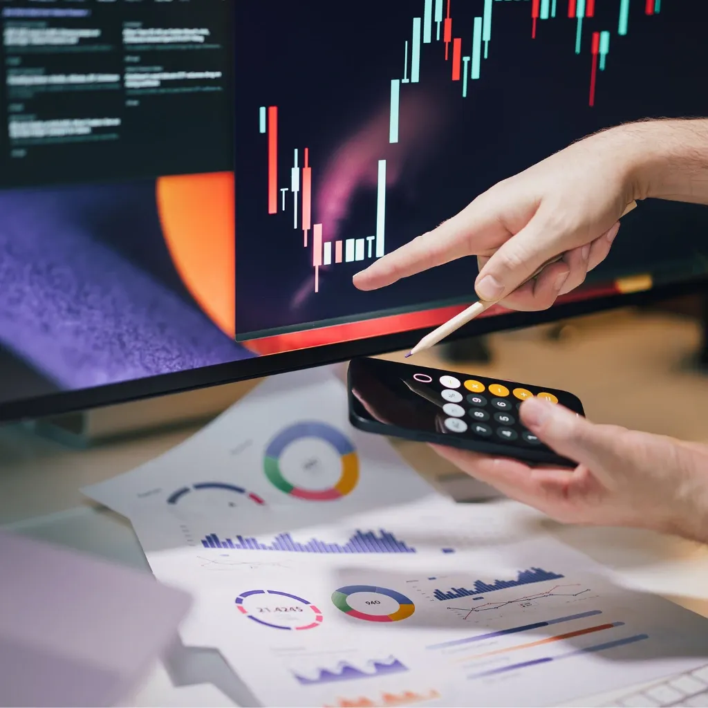 Person analyzes financial charts on a monitor, supplemented by charts and calculations on the desk.