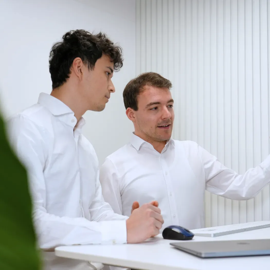Two colleagues in white shirts discuss something on a screen while seated at a desk with a laptop and mouse.