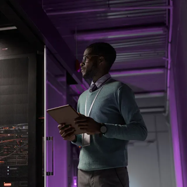 IT-Mitarbeiter mit Tablet steht vor einem Serverrack im violetten Licht.
