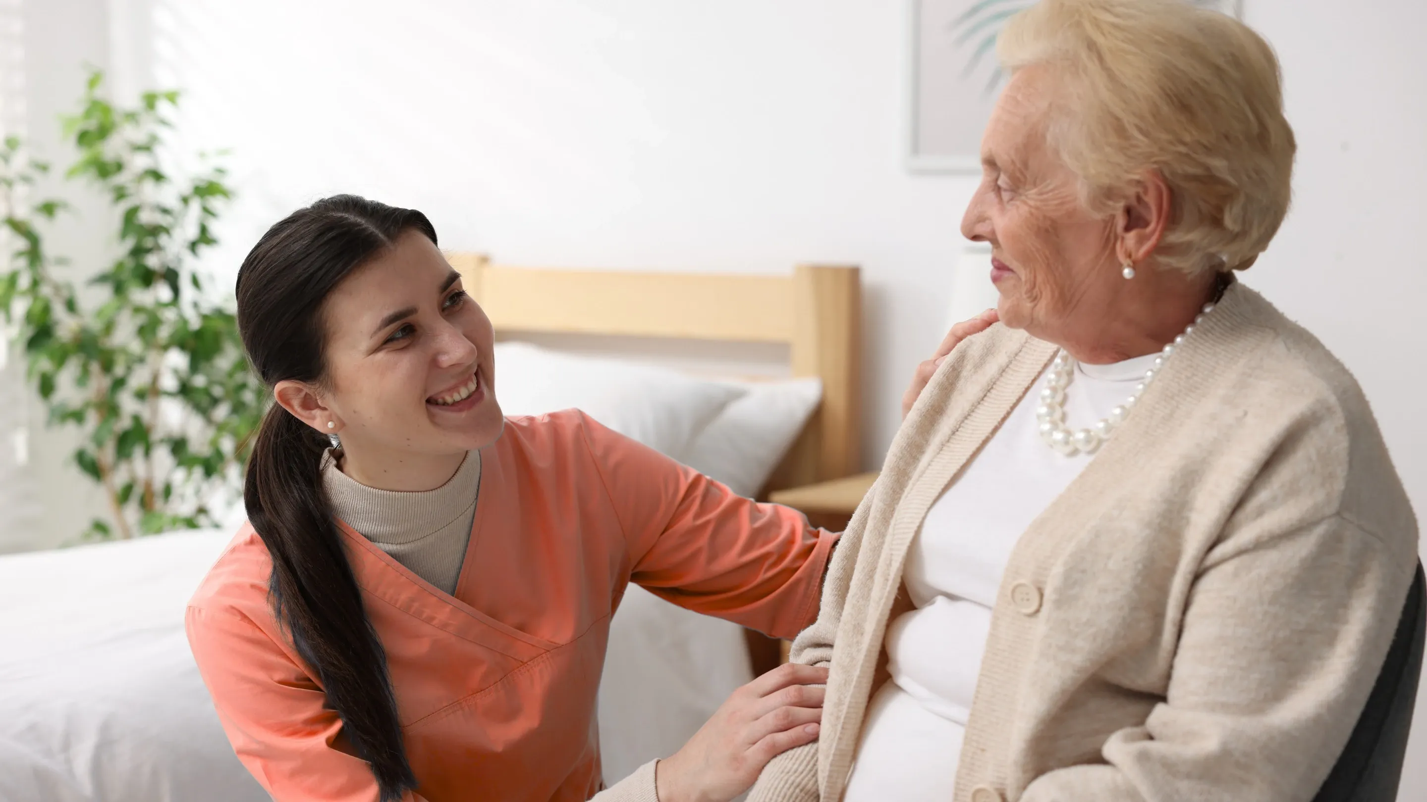 Eine junge Pflegerin in rosa Kleidung lächelt eine ältere Frau an.