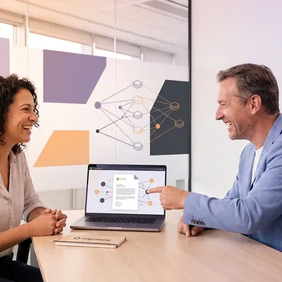 Two smiling professionals discussing a network diagram on a laptop, OriginStamp notebook on desk.