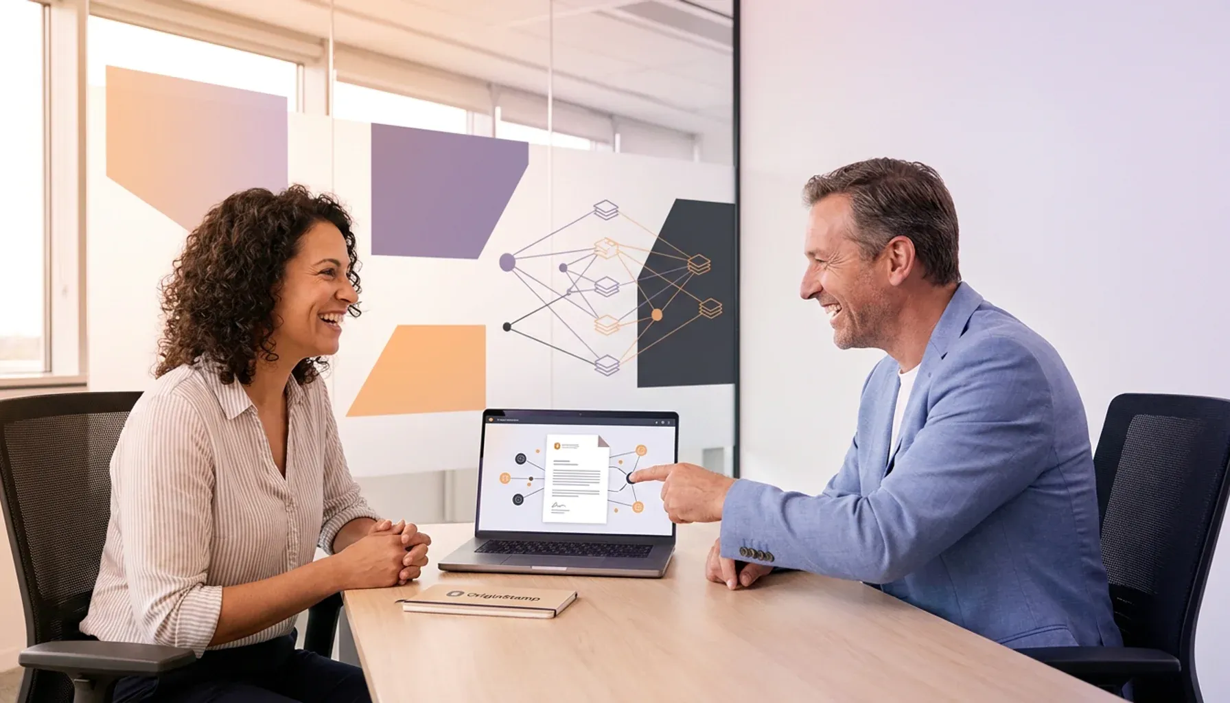 Two smiling professionals discussing a network diagram on a laptop, OriginStamp notebook on desk.