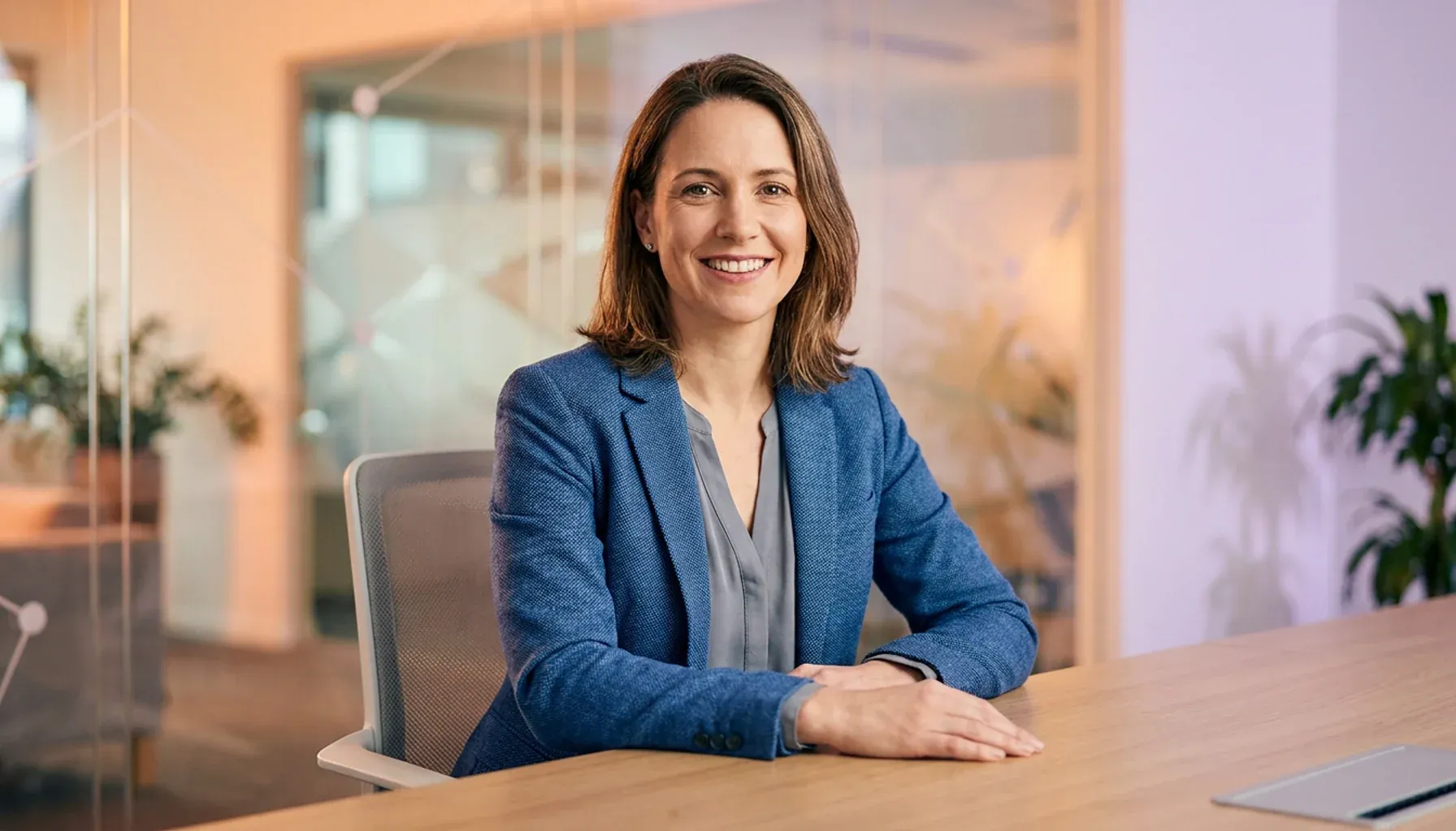Smiling businesswoman in a blue blazer sitting at a desk in a modern office.
