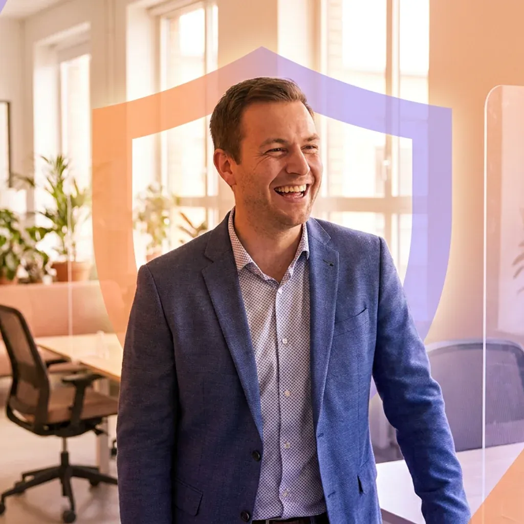 Smiling businessman talking to a colleague in an office with a shield graphic overlay.