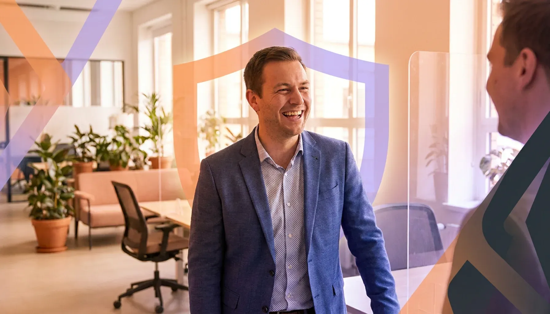Smiling businessman talking to a colleague in an office with a shield graphic overlay.