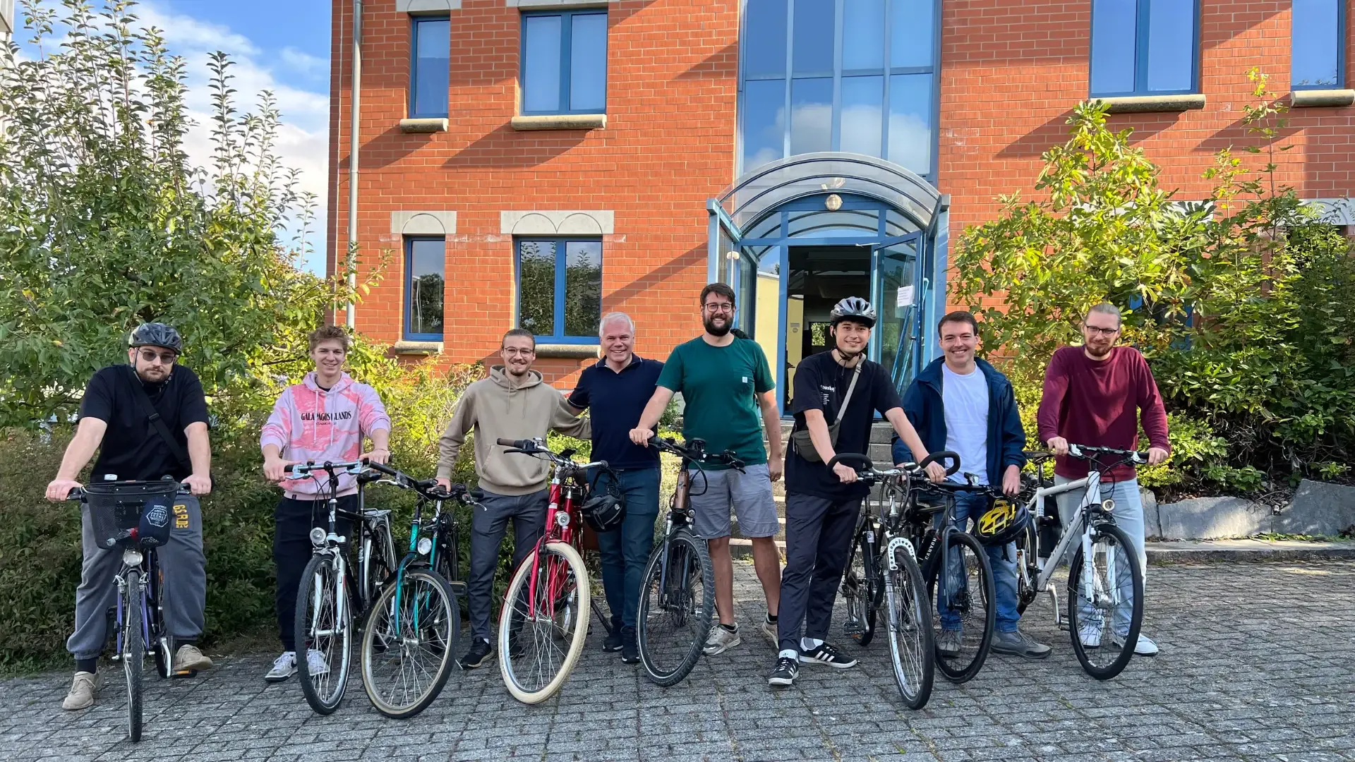 Acht Männer mit Fahrrädern posieren vor einem Backsteingebäude.