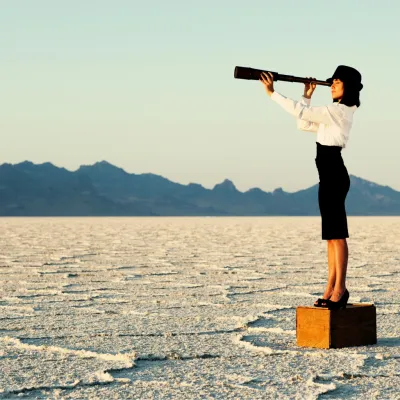Woman standing on a box in a vast desert landscape, looking through a telescope toward distant mountains.