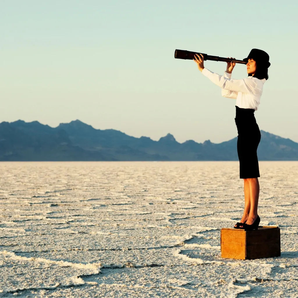 Woman standing on a box in a vast desert landscape, looking through a telescope toward distant mountains.