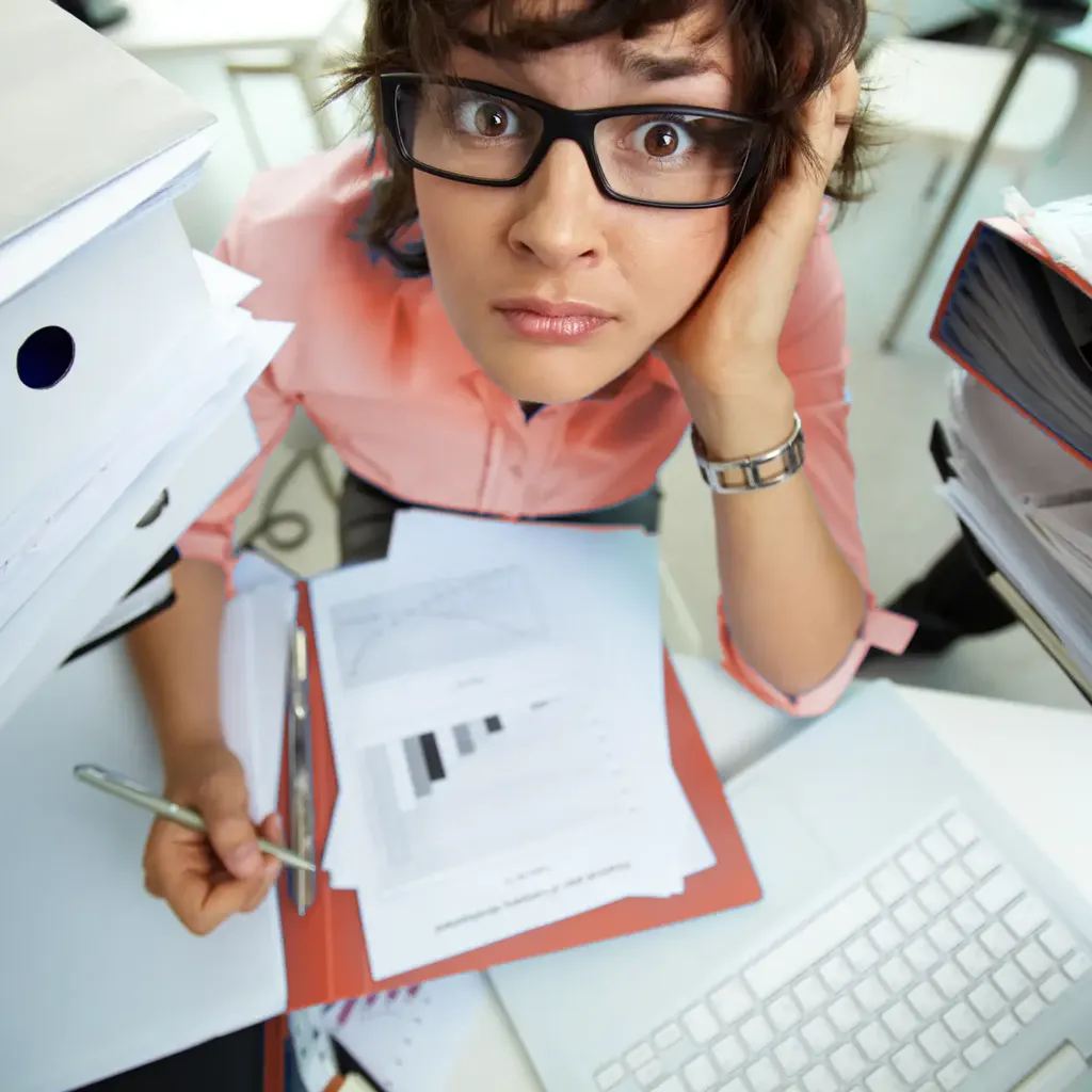 Gestresste Büroangestellte sitzt zwischen Aktenstapeln und Dokumenten am Schreibtisch und blickt überfordert in die Kamera
