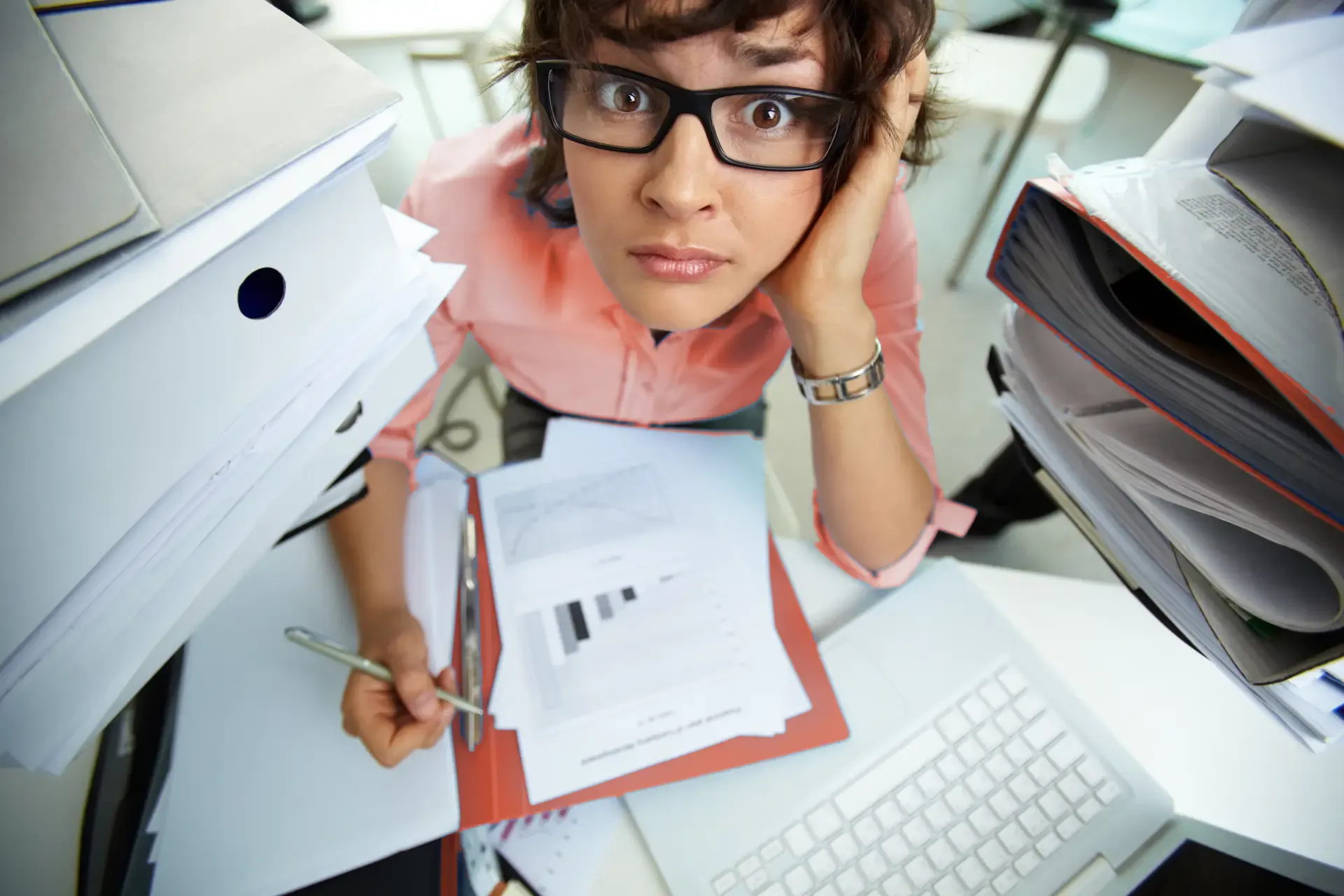Office worker sitting between tall stacks of folders at a desk, holding a pen and looking into the camera.