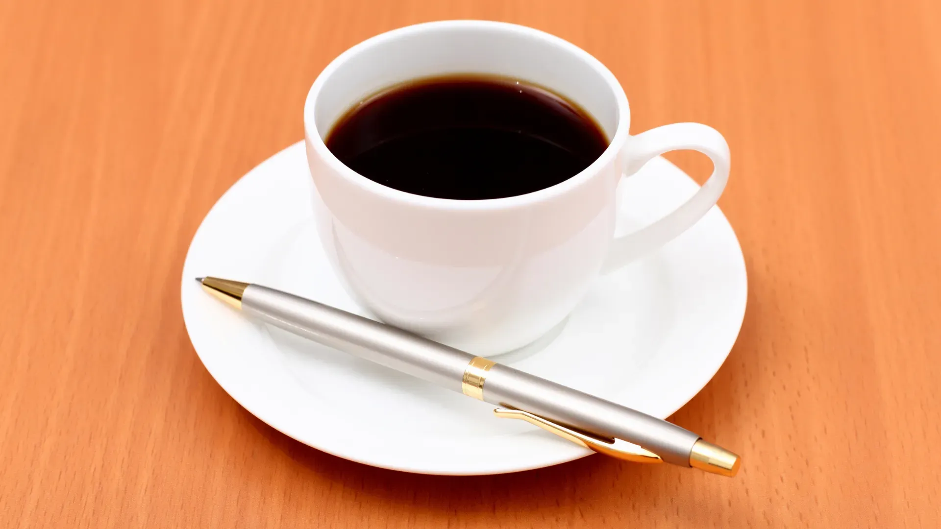Cup of black coffee on saucer with ballpoint pen next to it.